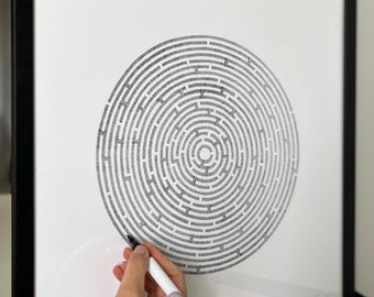 May include: Black framed artwork featuring a detailed, circular maze design in black ink on white paper. A hand holding a white marker is positioned near the maze, suggesting the artwork is interactive or customizable. The frame is black.