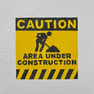 May include: A square yellow and black caution sign featuring the text CAUTION at the top and AREA UNDER CONSTRUCTION at the bottom. In the center, a black silhouette icon depicts a person using a shovel to dig into a pile of dirt.