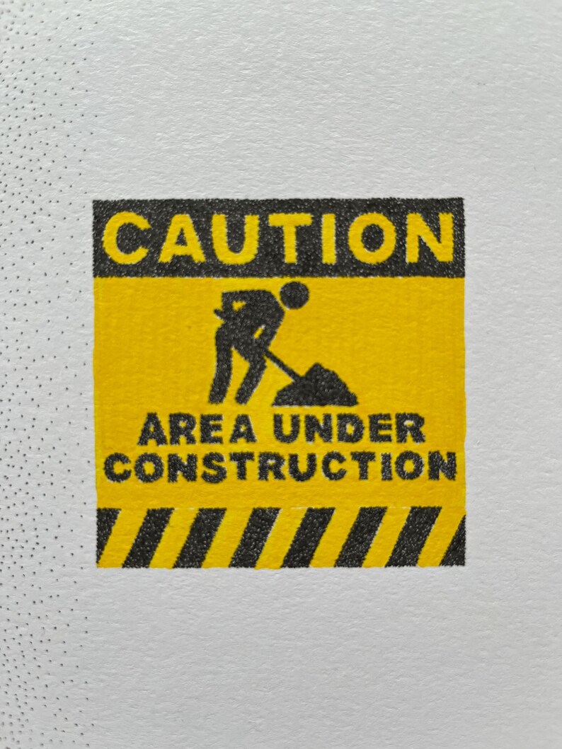 May include: A square yellow and black caution sign featuring the text CAUTION at the top and AREA UNDER CONSTRUCTION at the bottom. In the center, a black silhouette icon depicts a person using a shovel to dig into a pile of dirt.