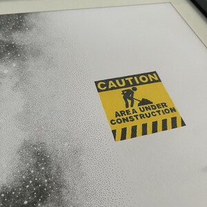 May include: A framed print featuring a stippled black and white galaxy design on a white background. A yellow square caution sign with black text reads CAUTION AREA UNDER CONSTRUCTION and includes a graphic of a person digging with a shovel.