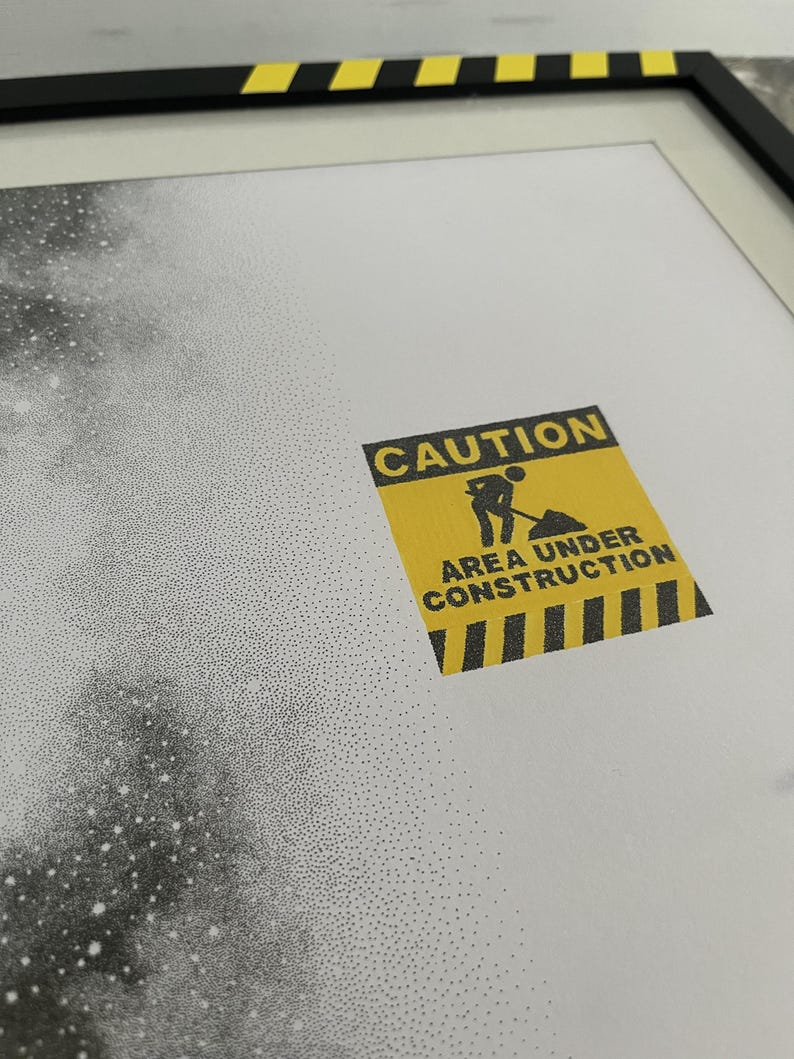 May include: A framed print featuring a stippled black and white galaxy design on a white background. A yellow square caution sign with black text reads CAUTION AREA UNDER CONSTRUCTION and includes a graphic of a person digging with a shovel.
