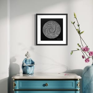 May include: A square black and white framed wall art print featuring a circular spiral pattern composed of tiny white dots on a solid black background, displayed in a black frame with a white mat border, hanging on a plain white wall.