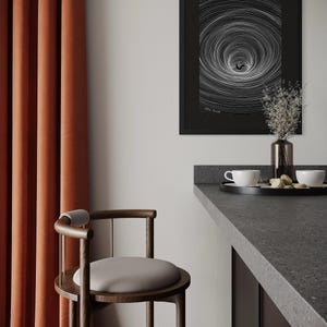 May include: A framed black and white print featuring a swirling vortex pattern with a small figure at the center, hanging on a wall above a dark gray counter with a black tray, two white cups, and a glass vase containing dried flowers.