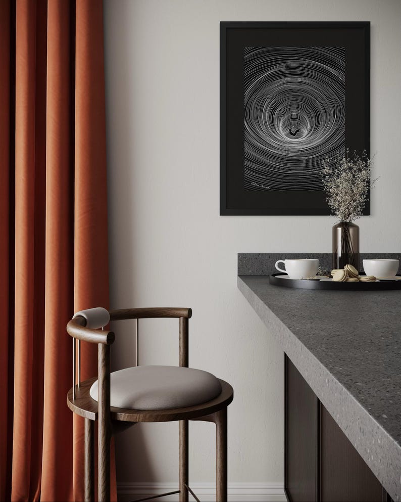May include: A framed black and white print featuring a swirling vortex pattern with a small figure at the center, hanging on a wall above a dark gray counter with a black tray, two white cups, and a glass vase containing dried flowers.