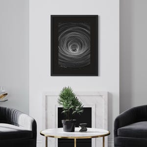 May include: A framed black and white art print featuring a central dark silhouette surrounded by concentric circular lines that create a tunnel-like effect. The artwork is enclosed in a thin black frame with a dark mat, displaying a signature at the bottom.