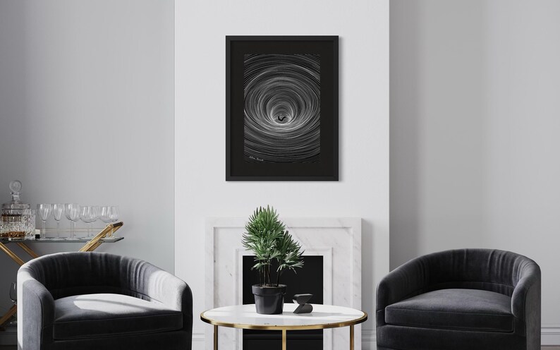 May include: A framed black and white art print featuring a central dark silhouette surrounded by concentric circular lines that create a tunnel-like effect. The artwork is enclosed in a thin black frame with a dark mat, displaying a signature at the bottom.