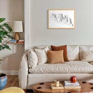 May include: A framed black and white line drawing of a mountain range hanging on a light grey wall above a cream-coloured sofa with various throw pillows, a wooden coffee table, and a lamp on a small shelf.