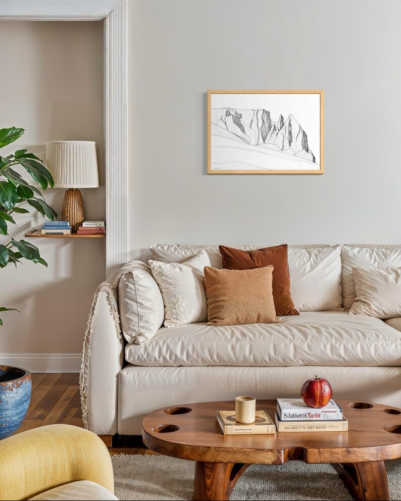 May include: A framed black and white line drawing of a mountain range hanging on a light grey wall above a cream-coloured sofa with various throw pillows, a wooden coffee table, and a lamp on a small shelf.