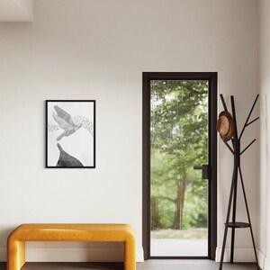 K&ouml;nnte beinhalten: Ein rechteckiger, schwarz gerahmter Kunstdruck mit einer monochromen Illustration eines fliegenden Vogels &uuml;ber einer dunklen Form, platziert &uuml;ber einer senfgelben Polsterbank neben einem dunkelbraunen Garderobenst&auml;nder mit Hut.