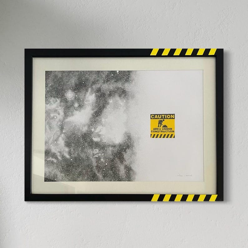 May include: A framed print featuring a black and white nebula on the left and a white background on the right. A yellow caution sign with the text CAUTION AREA UNDER CONSTRUCTION is centered on the right. The black frame has yellow and black stripes.