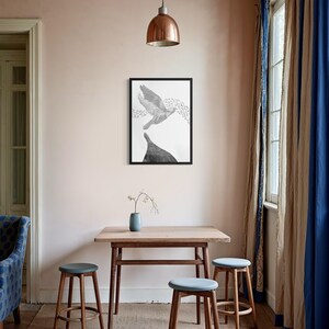 K&ouml;nnte beinhalten: Ein gerahmter schwarz-wei&szlig;er Wandkunstdruck, der einen fliegenden Vogel &uuml;ber einer dunklen, massiven Form zeigt, pr&auml;sentiert an einer hellrosa Wand &uuml;ber einem Holztisch mit drei Hockern und einer kleinen blauen Vase.