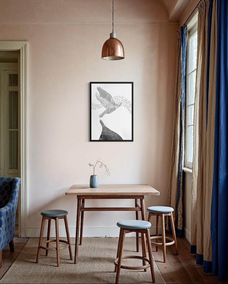 K&ouml;nnte beinhalten: Ein gerahmter schwarz-wei&szlig;er Wandkunstdruck, der einen fliegenden Vogel &uuml;ber einer dunklen, massiven Form zeigt, pr&auml;sentiert an einer hellrosa Wand &uuml;ber einem Holztisch mit drei Hockern und einer kleinen blauen Vase.