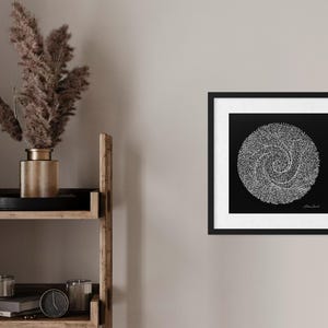 May include: A square framed wall art piece featuring a black background with a white circular spiral design composed of numerous small dots. The artwork is enclosed in a thin black frame with a white mat border, displaying a signature in the corner.