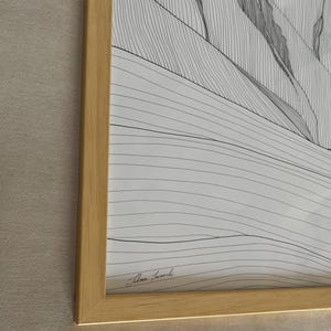 May include: A framed black and white line art print featuring a landscape of rolling hills and mountains. The artwork is enclosed in a light-coloured wooden frame. A signature is visible in the bottom left corner of the white paper print.