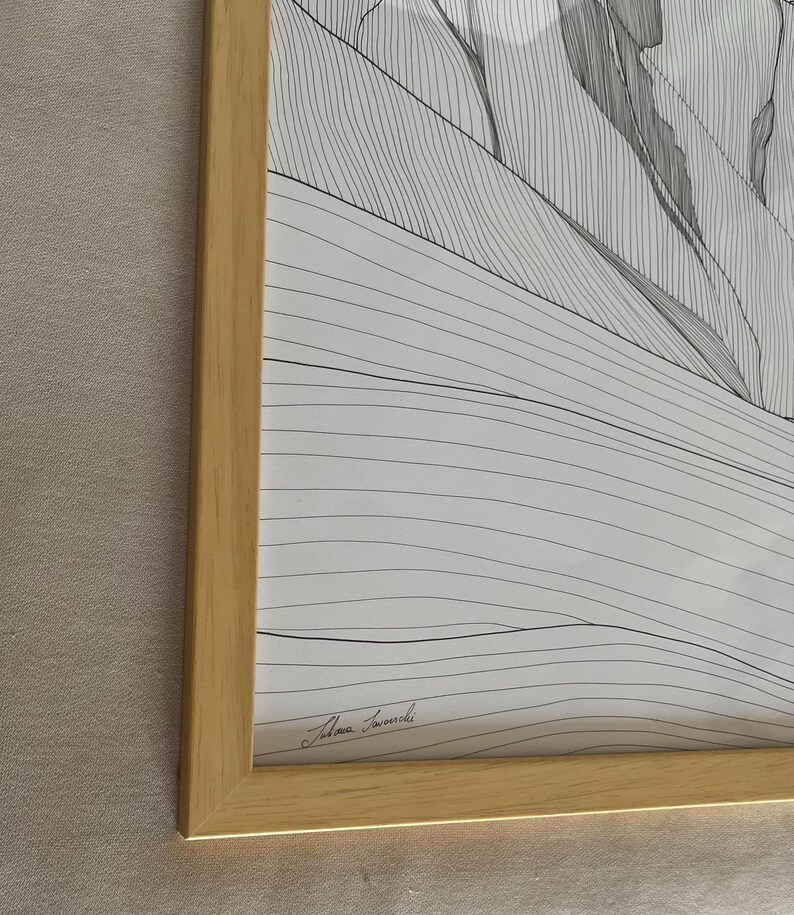 May include: A framed black and white line art print featuring a landscape of rolling hills and mountains. The artwork is enclosed in a light-coloured wooden frame. A signature is visible in the bottom left corner of the white paper print.