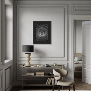 May include: A framed black and white wall art print featuring a circular vortex pattern with radiating lines. The artwork is displayed in a thin black frame and is mounted on a light gray wall above a wooden console table with books and a lamp.