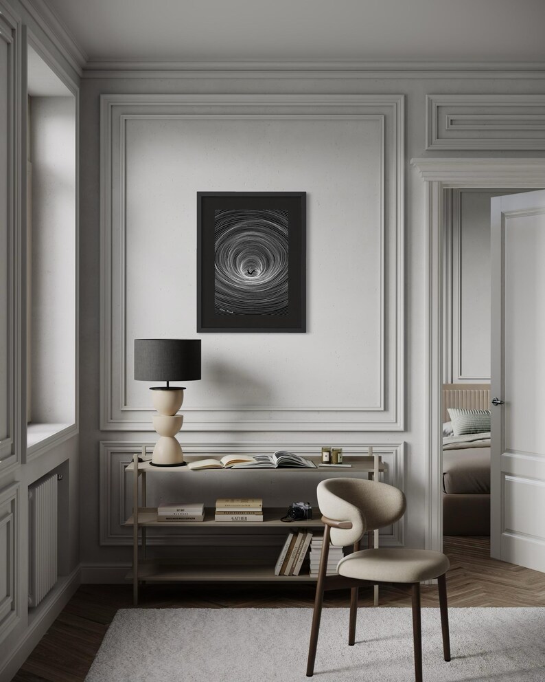 May include: A framed black and white wall art print featuring a circular vortex pattern with radiating lines. The artwork is displayed in a thin black frame and is mounted on a light gray wall above a wooden console table with books and a lamp.