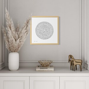 May include: A square framed wall art piece featuring a black and white circular maze design on a white background. The frame is a light wood tone. The artwork is displayed on a wall, centred above a white cabinet with decorative objects.