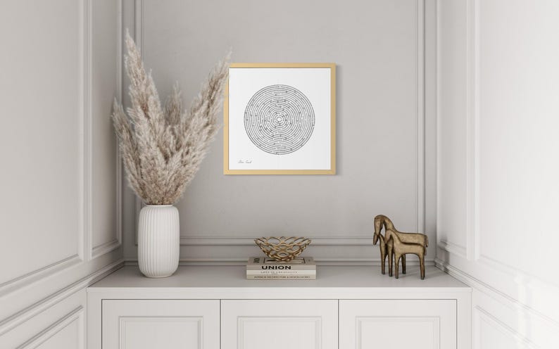 May include: A square framed wall art piece featuring a black and white circular maze design on a white background. The frame is a light wood tone. The artwork is displayed on a wall, centred above a white cabinet with decorative objects.