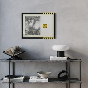 May include: A rectangular wall art print in a black frame with yellow and black hazard stripes on the top right and bottom left corners. The print features a black and white textured abstract design and a yellow caution sign with text.