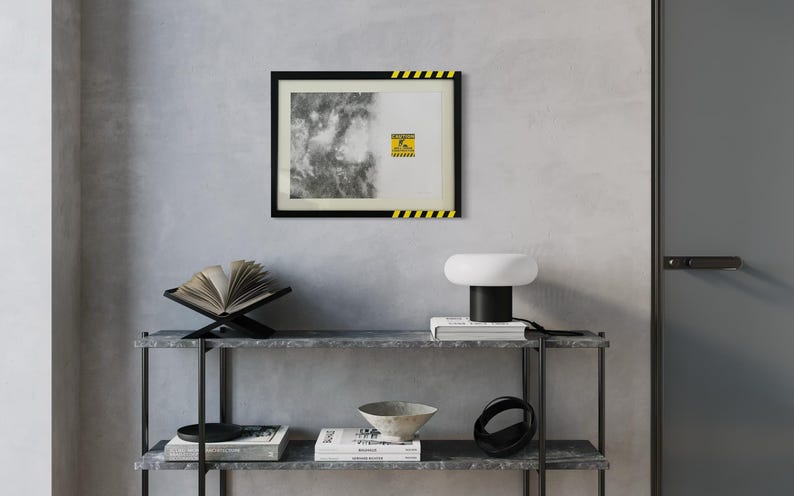 May include: A rectangular wall art print in a black frame with yellow and black hazard stripes on the top right and bottom left corners. The print features a black and white textured abstract design and a yellow caution sign with text.