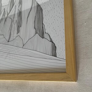 May include: A framed black and white line art print featuring a mountain landscape with jagged peaks and a stippled sky. The artwork is encased in a light-toned wooden frame with a mitered corner design, showcasing detailed vertical line work.