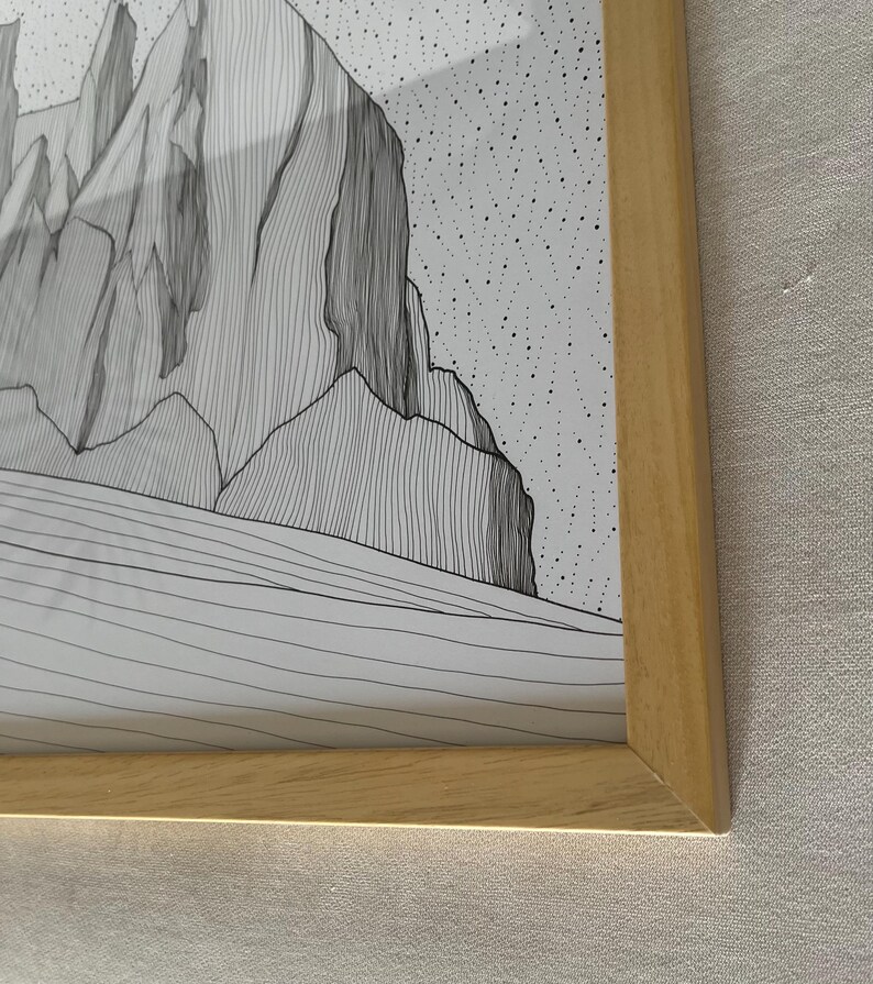May include: A framed black and white line art print featuring a mountain landscape with jagged peaks and a stippled sky. The artwork is encased in a light-toned wooden frame with a mitered corner design, showcasing detailed vertical line work.