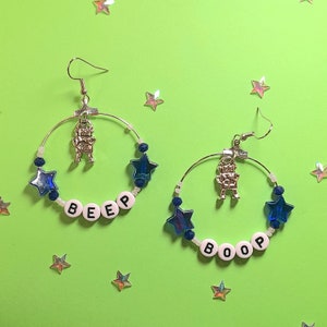 May include: A pair of hoop earrings with silver-tone metal hoops. Each earring features a small silver robot charm, blue star-shaped beads, and white letter beads spelling "BEEP" and "BOOP". The background is a bright green with star-shaped confetti.