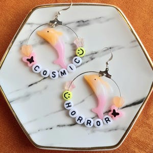 May include: A pair of hoop earrings with a dolphin charm, the dolphin is gradient orange to pink. The hoops have the words "COSMIC HORROR" spelled out with white letter beads. Additional charms include butterflies, stars, and smiley faces.