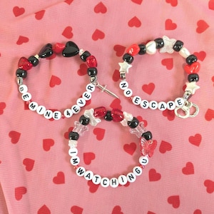 May include: Three beaded bracelets with red, black, and white beads. One bracelet says "BE MINE 4EVER", another says "NO ESCAPE", and the third says "I'M WATCHING U". The bracelets have heart, star, and butterfly charms.