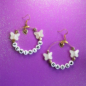 May include: A pair of gold hoop earrings with white butterfly and star charms. One earring spells "FEMININE" in white letter beads, the other spells "RAGE". The earrings have gold heart charms and are set against a purple background.