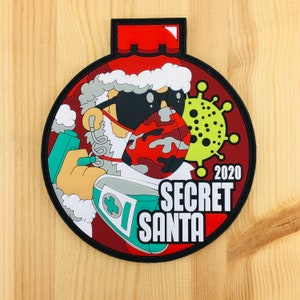 Può includere: Un ornamento natalizio rosso e bianco con un Babbo Natale dei cartoni animati che indossa una maschera facciale mimetica e tiene una bottiglia di disinfettante per le mani. L'ornamento presenta anche una grafica di coronavirus verde e gialla e il testo "2020 Secret Santa".
