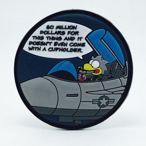 May include: A round, blue and gray patch with a cartoon bird in a fighter jet. The bird is holding a drink and a speech bubble above the jet says "60 million dollars for this thing and it doesn't even come with a cupholder."
