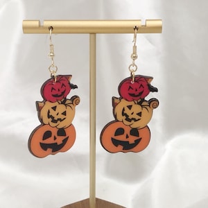 May include: A pair of Halloween-themed drop earrings. Each earring features three stacked pumpkins in orange, yellow, and red, with carved faces. The earrings hang from gold-coloured hooks, ideal for festive occasions.