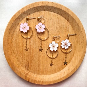 May include: Three pairs of gold dangle earrings with a flower charm and a star charm. The flowers are pink and white with gold accents. The earrings are on a wooden surface.