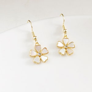 May include: A pair of gold-coloured flower earrings. Each earring features a five-petal flower design with a white and pink mother-of-pearl effect and a small clear crystal in the centre. The earrings have a hook closure.