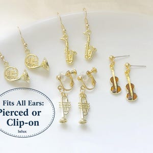 Peut inclure: Boucles d'oreilles dorées en forme d'instruments de musique : cors, saxophones, trompettes et violons. Certaines sont de type clip. Le texte de l'image indique : "Convient à toutes les oreilles : Percées ou à clip".