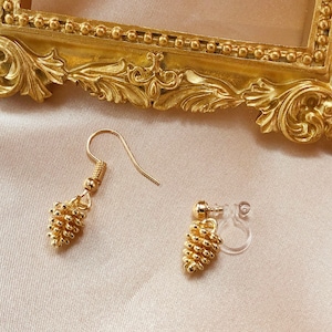 Puede incluir: Pendientes dorados con un diseño texturizado en forma de piña. Un pendiente tiene un gancho y el otro tiene un respaldo transparente con clip. Los pendientes se exhiben sobre una superficie suave y clara, con un marco dorado decorativo en el fondo.