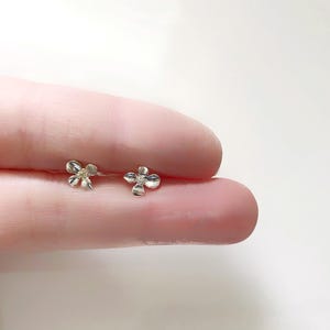 May include: A pair of small, silver flower-shaped stud earrings. Each earring has a four-petal design with a slightly textured surface. The earrings are displayed on a light pink background.