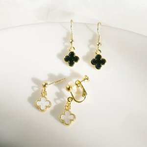 Op de afbeelding: Een paar gouden oorbellen met zwarte en witte klavertjevier-charms. De oorbellen zijn van het hangende type met een oorbelsteker en een clip-sluiting.