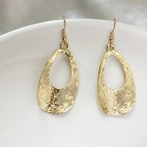 May include: A pair of gold-coloured teardrop earrings. The earrings have a textured surface with a fingerprint-like pattern. The earrings hang from gold-coloured fish hook ear wires. The earrings are a jewellery accessory.