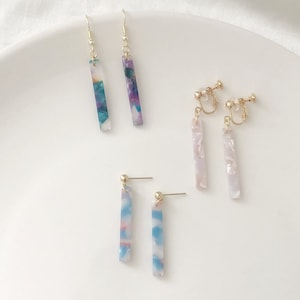 May include: Four pairs of earrings with long, rectangular, colourful, patterned, acrylic dangles. Two pairs are gold hook earrings and two pairs are gold clip-on earrings.