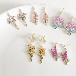 Könnte beinhalten: Sortiment an floralen Ohrringen in Gold mit Emaille-Details. Die Ohrringe zeigen verschiedene Blumendesigns, darunter Hortensien, Lavendel und Iris. Einige Ohrringe sind im Clip-Stil.