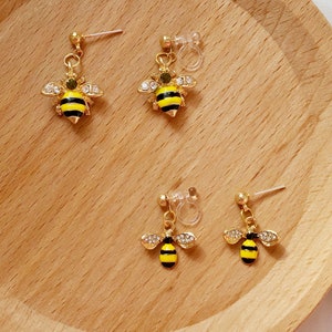Peut inclure: Une paire de boucles d'oreilles en forme d'abeille dorées avec des rayures noires et jaunes et des strass transparents. Les boucles d'oreilles ont un fermoir à clip.