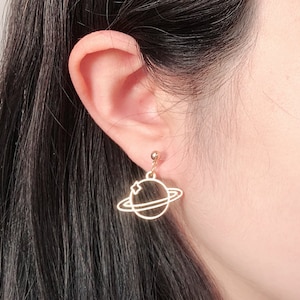 May include: Gold-colored planet earrings. Each earring features a delicate outline of a planet with a star, suspended from a small gold ball. The earrings are designed to dangle, adding a touch of celestial charm.