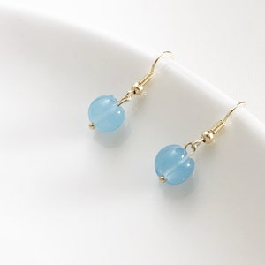 May include: A pair of earrings featuring light blue, round beads. The beads are suspended from gold-colored hooks. The earrings are displayed on a white surface, showcasing their delicate design and color.