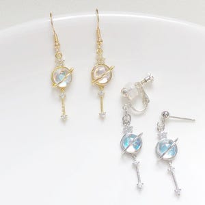 Puede incluir: Pendientes con temática celestial. Dos pares de pendientes colgantes, uno dorado y otro plateado, presentan un diseño de planeta con un orbe iridiscente y brillante y un acento de cristal colgante. Un par tiene cierres de clip.