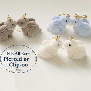 May include: Earrings shaped like bunnies in grey, pale blue, and white. Each pair has a gold-coloured ear wire. The image includes text that reads "Fits All Ears: Pierced or Clip-on".