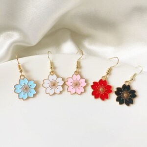 May include: Five pairs of gold-tone earrings with a cherry blossom design. The earrings are available in blue, white, pink, red, and black.
