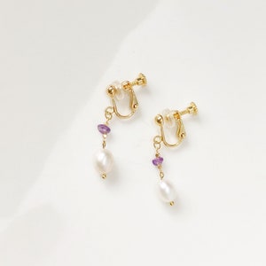 May include: A pair of clip-on earrings featuring gold-colored metal hardware. Each earring has a small cluster of purple gemstones and a white, teardrop-shaped pearl. The earrings are displayed against a white background.